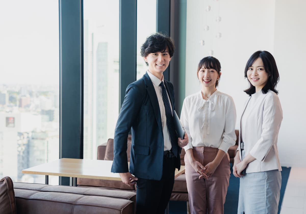 高層オフィスの窓際に立つ3人の若手ビジネスパーソン。紺のスーツを着た男性と、白いブラウスにピンクベージュのパンツを着た女性、白いジャケットの女性が並んで微笑んでいる。背景には都市の景観が広がる。