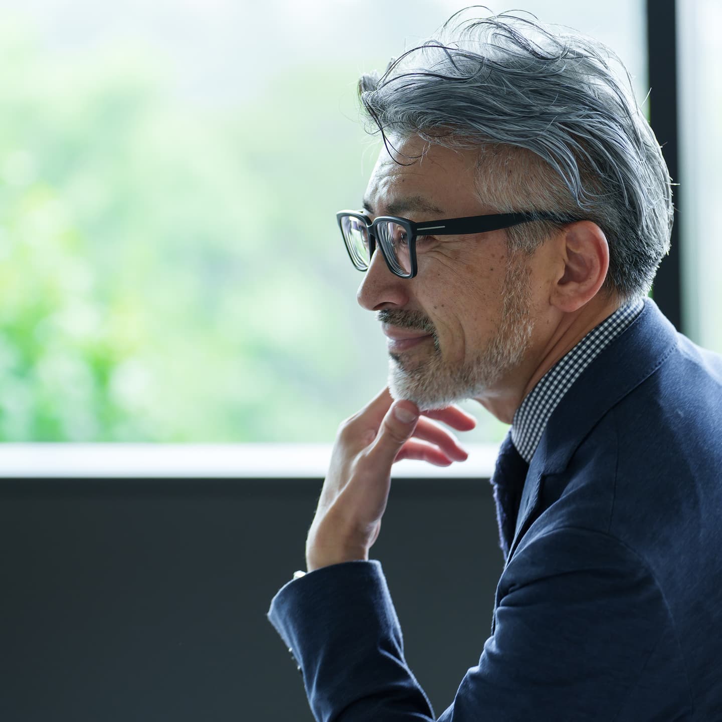 窓際で思慮深い表情を見せる眼鏡をかけた白髪交じりの中年男性経営者。手を顎に当て、遠くを見つめている様子