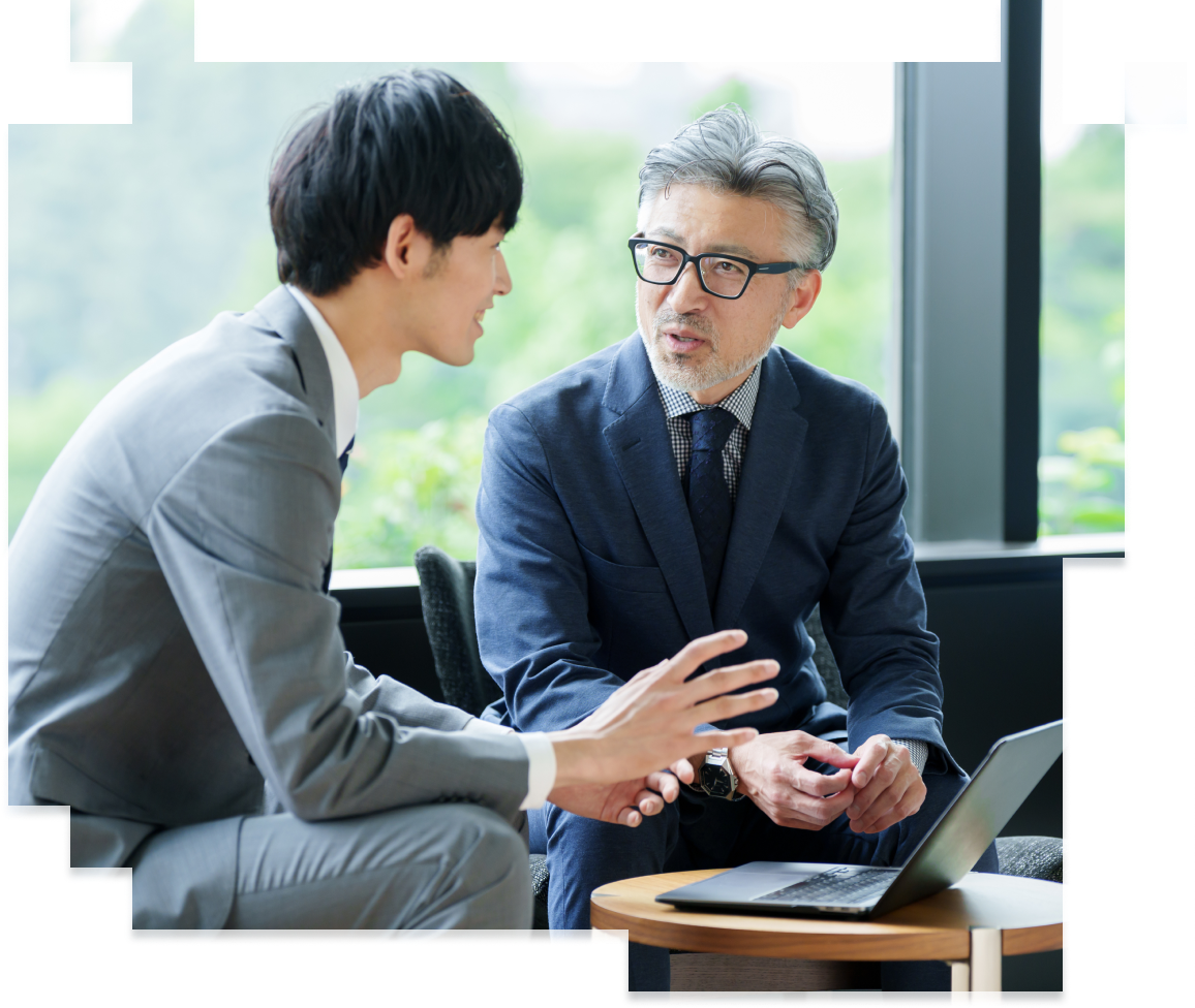 窓際のミーティングスペースで対話する年齢の異なるビジネスマン。若手のグレースーツの男性がジェスチャーを交えて説明し、紺のスーツに眼鏡をかけた白髪の上司が真剣に聞いている。丸テーブルにはノートパソコンがある