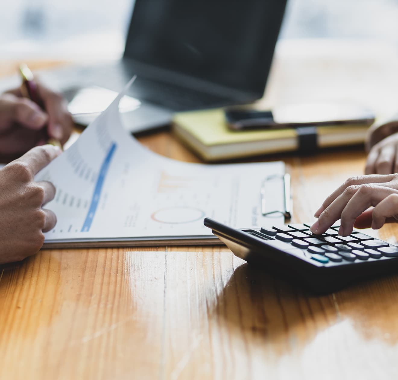 木製テーブル上での会計作業。グラフ付き書類を確認する手と電卓を操作する手。背景にはぼやけたノートパソコンとメモ帳が見える