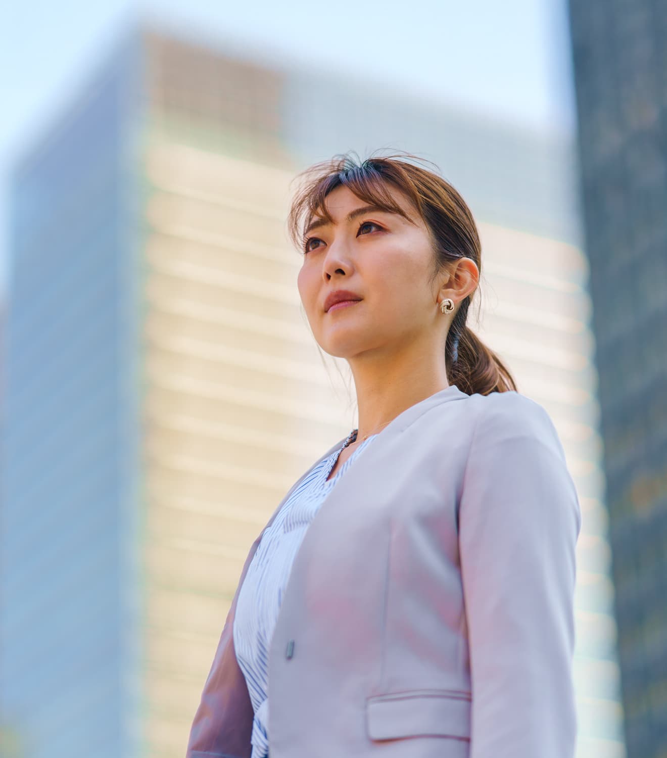 高層ビルを背景に上を見上げる淡いピンク色のジャケットを着た女性ビジネスパーソン。ポニーテールの髪型で、前向きで自信に満ちた表情をしている