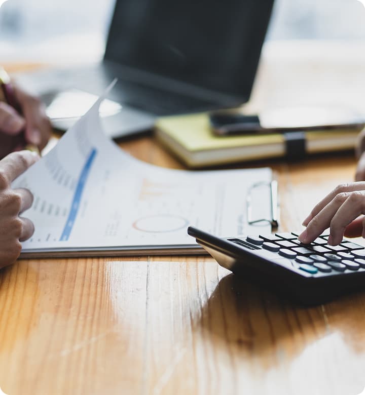 木製テーブル上での会計作業。グラフ付き書類を確認する手と電卓を操作する手。背景にはぼやけたノートパソコンとメモ帳が見える