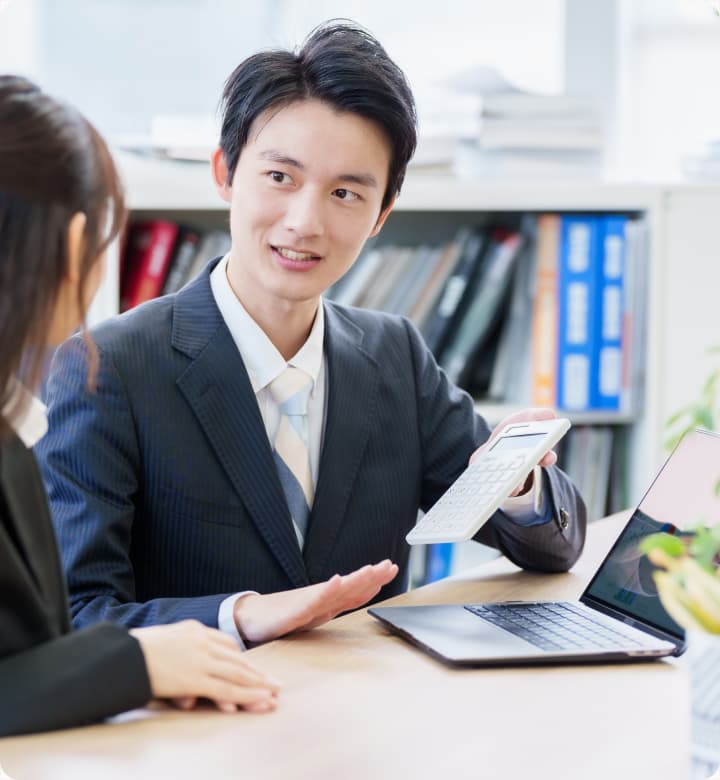 オフィスで電卓を手に会話するストライプスーツの若いビジネスマン。ノートパソコンの前で相手に説明している様子。背景には本棚がある