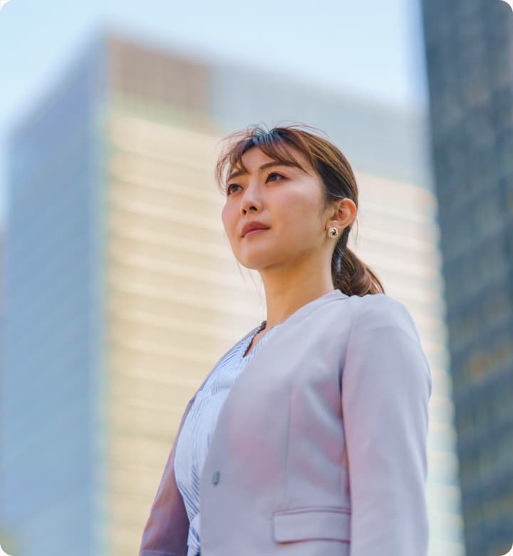 高層ビルを背景に上を見上げる淡いピンク色のジャケットを着た女性ビジネスパーソン。ポニーテールの髪型で、前向きで自信に満ちた表情をしている