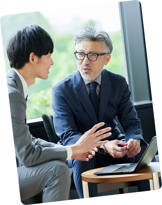 窓際のテーブルで対話する2人のビジネスマン。グレーのスーツを着た若い男性と、眼鏡をかけた白髪交じりの中年男性が真剣に会話している。テーブルにはノートパソコンが置かれている。