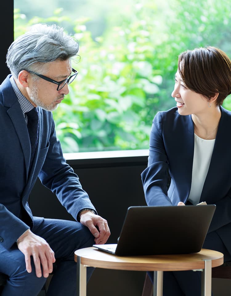 オフィスの窓際で、ノートパソコンを前に会話をする紺色のスーツを着た男性と女性のビジネスパーソン。背景には緑の木々が見える。