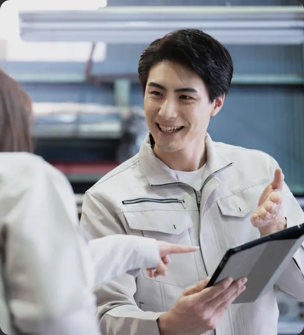 工場で白い作業服を着た若い男性がタブレットを持ちながら同僚と笑顔で会話している様子。産業施設の背景が見える。