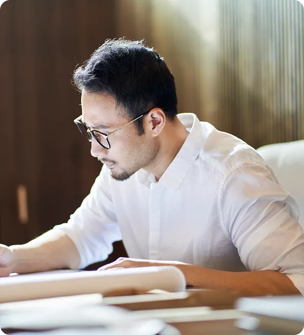 眼鏡をかけた白いシャツの男性が机で書類を確認している様子。集中して資料に目を通している。