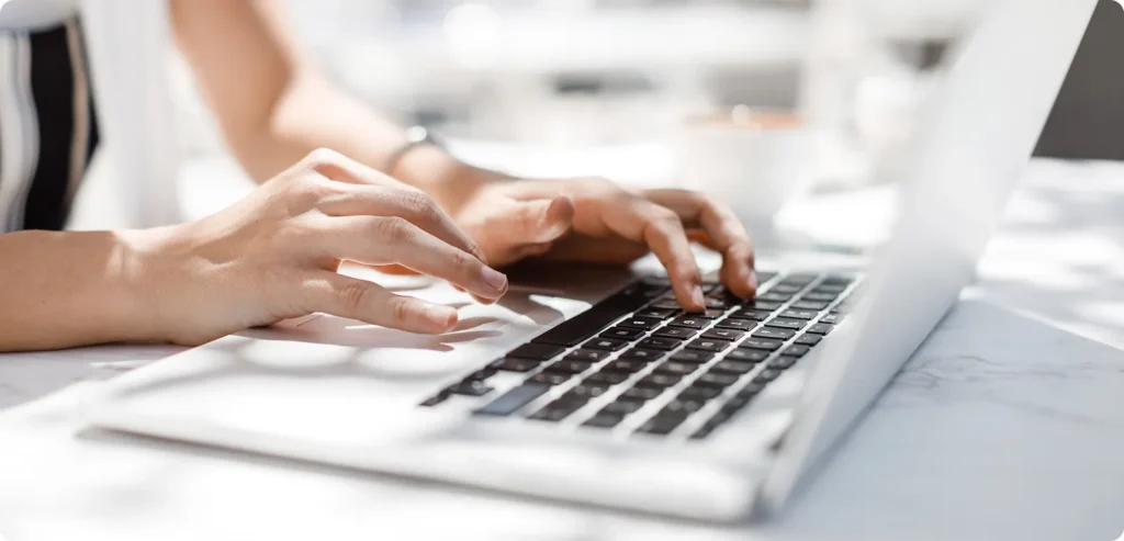 白い机でノートパソコンのキーボードをタイピングする手元のクローズアップ。明るいオフィス環境での作業風景。