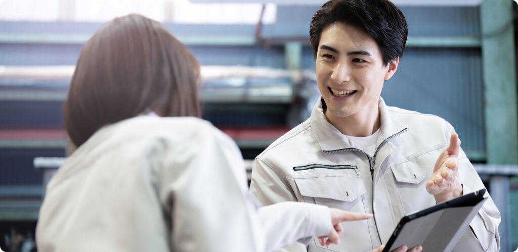 工場で白い作業服を着た若い男性がタブレットを持ちながら同僚と笑顔で会話している様子。産業施設の背景が見える。